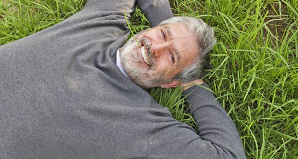 Mann in den besten Jahren mit Bart, grauen Haaren und grauem Pullover liegt mit dem Rücken im Gras und hat einen Grashalm zwischen den Zähnen